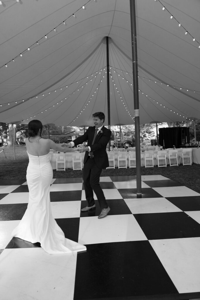 Private Estate Tented Wedding ~ Lyme, CT - Gambo Photo Co. - a black and white photo featuring the bride and groom having their private first dance on a black and white checked dance floor under a sailcloth high peak tent with bistro lighting. 