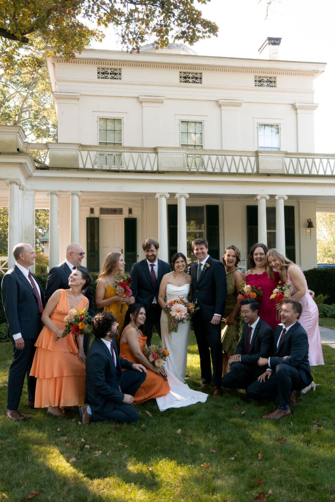 Private Estate Tented Wedding ~ Lyme, CT - Gambo Photo Co. - the wedding party dressed in navy blue suits and the bridal party in gray of bright orange, pink, yellow, and green summer dresses posing infant of the "The Big House" at Griswold Point in Lyme, CT 