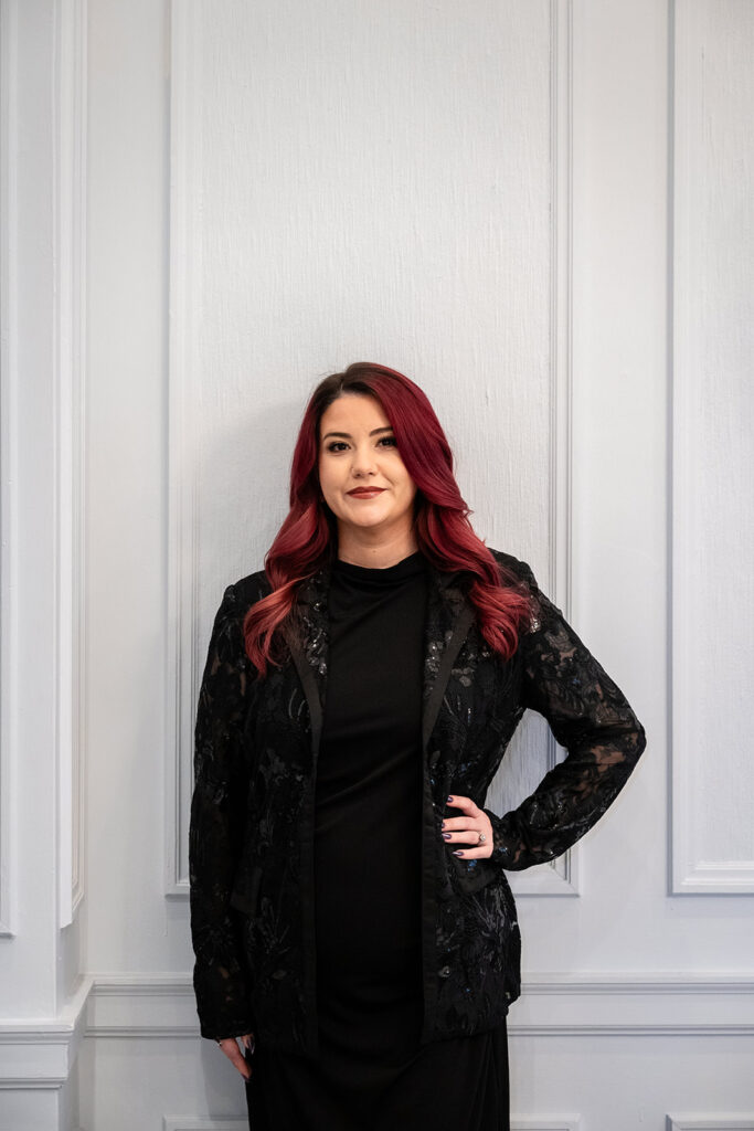 Young Women with red hair standing in front of a white wall with crown molding wearing a black blazer adorned with lace and sequence and black dress. J. Parsons Photography 