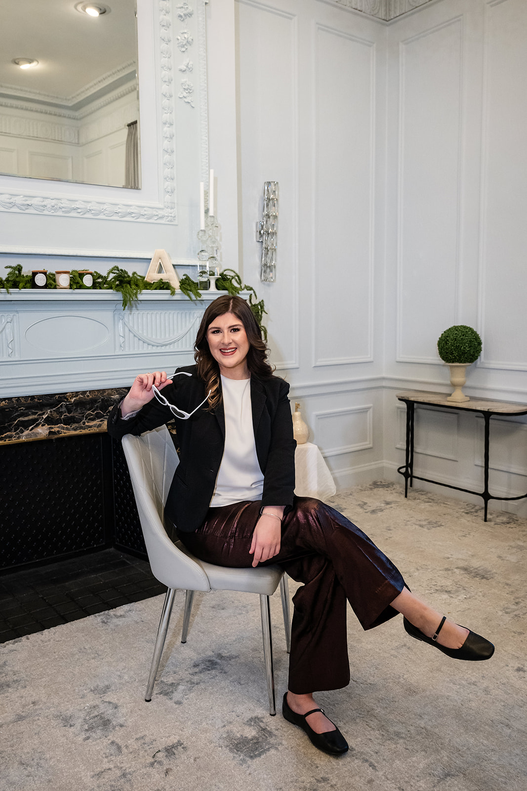 Tall Brunette Girl sitting in a chair in front of a fireplace dressed with greenery, holding a pair a white heart shaped sunglasses. J. Parsons Photography 