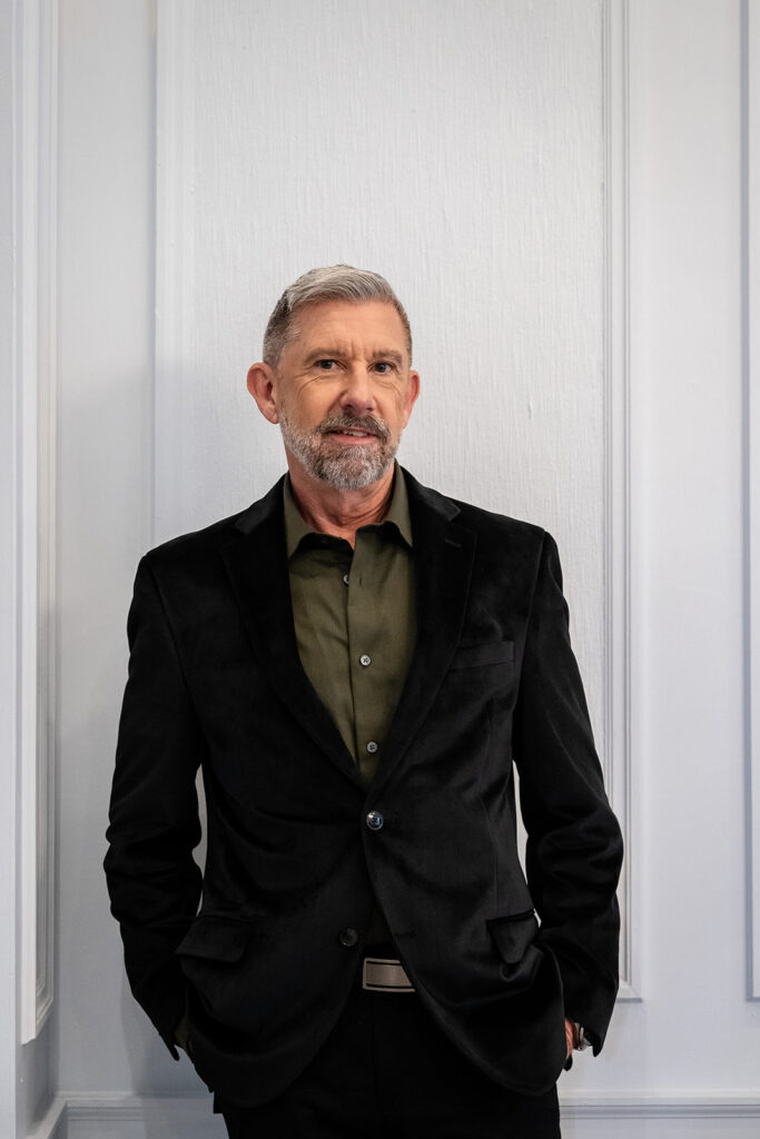 Older Gentleman standing in front of a white wall with crown molding earring a black velvet jacket and an olive green shirt. J. Parsons Photography 